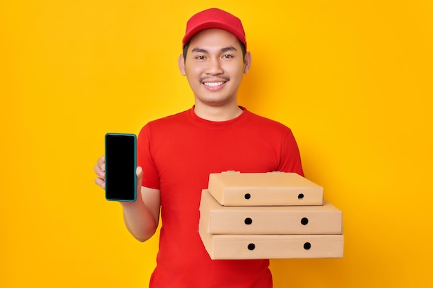 Enthousiaste jeune homme asiatique en casquette rouge tshirt uniforme livreur employé travailler comme concessionnaire courrier tenir boîte à pizza montrant un téléphone portable à écran blanc isolé sur fond jaune Concept de livraison de pizza