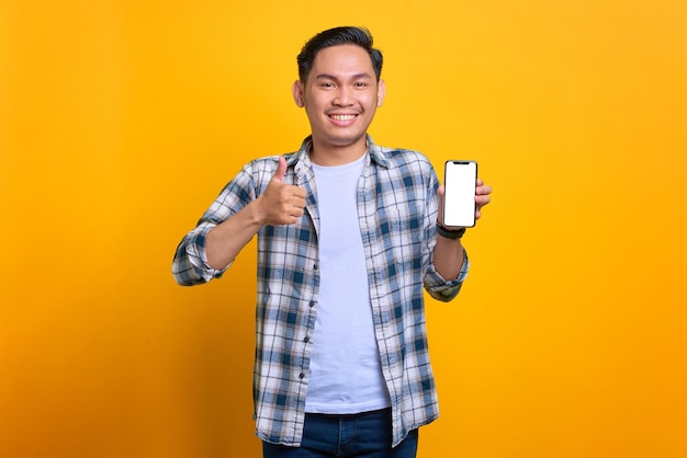 Enthousiaste jeune homme asiatique en chemise à carreaux tenant un téléphone portable avec écran blanc montrant le geste du pouce vers le haut isolé sur fond jaune