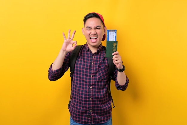 Enthousiaste jeune homme asiatique avec sac à dos détenant un passeport et des billets d'avion montrant un signe correct isolé sur fond jaune Tourisme et concept de voyage de vacances