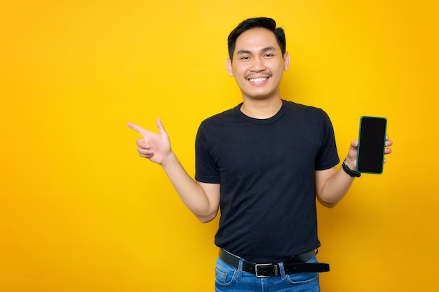 Enthousiaste jeune homme asiatique en t-shirt décontracté montrant un téléphone portable avec un écran blanc pointant le doigt de côté recommandant une application mobile isolée sur fond jaune Concept de style de vie des gens