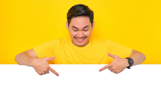 Enthousiaste jeune homme asiatique en t-shirt décontracté pointant vers le bas au panneau publicitaire blanc isolé sur fond jaune Concept de panneau d'affichage de promotion