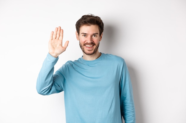 Enthousiaste jeune homme avec barbe disant bonjour, à la main amicale et en agitant pour vous saluer, debout sur fond blanc