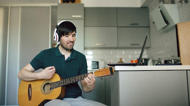 Enthousiaste jeune homme avec un casque assis à la cuisine apprenant à jouer de la guitare à l'aide d'un ordinateur portable