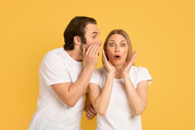 Enthousiaste jeune homme caucasien chuchotant à l'oreille d'une femme choquée en t-shirt blanc