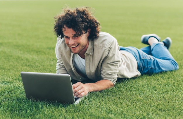 Enthousiaste jeune homme indépendant assis sur le sol travaillant sur un ordinateur portable Homme souriant aux cheveux bouclés utilisant un ordinateur portable pour discuter en ligne avec des amis sur l'herbe Homme tapant sur son ordinateur dans le parc