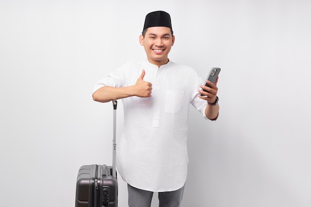 Enthousiaste jeune homme musulman asiatique debout tout en tenant une valise et un smartphone montrant le geste du pouce vers le haut isolé sur fond blanc Concept de Ramadan et eid Mubarak