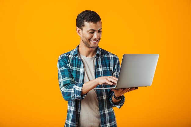 Enthousiaste jeune homme portant une chemise à carreaux debout isolé sur fond orange, tenant un ordinateur portable