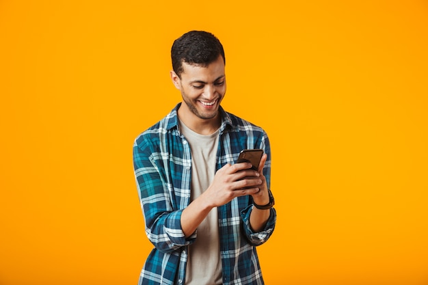 Enthousiaste jeune homme portant une chemise à carreaux debout isolé sur un mur orange, à l'aide de téléphone mobile