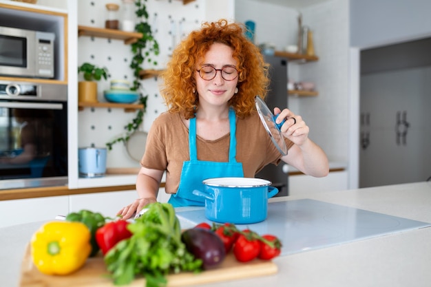 Enthousiaste jeune jolie femme en tablier préparer le déjeuner et sentir le plat dans la cuisine à la maison Femme préparant le dîner pour la famille à la nouvelle recette à la maison