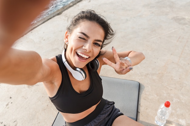 Enthousiaste jeune sportive au repos après l'entraînement à la plage, en prenant un selfie, de l'eau potable, assis sur un tapis de fitness