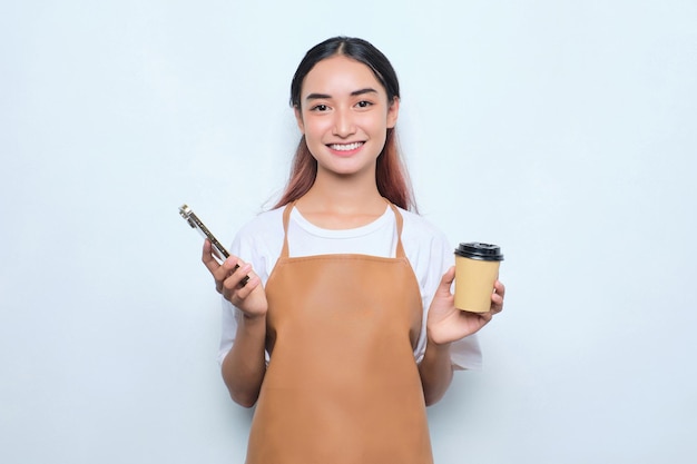 Enthousiaste jolie jeune fille barista en tablier tenant une tasse de café à emporter à l'aide d'un téléphone portable isolé sur fond blanc