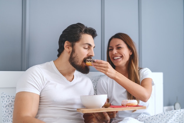 Enthousiaste magnifique brune caucasienne assise dans le lit le matin et nourrir son mari avec un beignet.