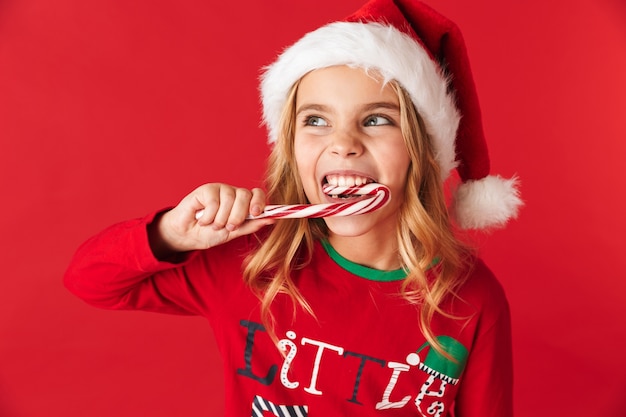 Enthousiaste petite fille portant le costume de Noël debout isolé, manger des bonbons de cône de bonbons de Noël