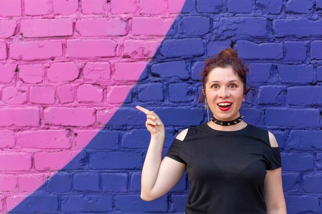 Enthousiaste positive jeune femme en vêtements goth posant contre un mur de briques bleu-violet avec copyspace. Le concept de contenu publicitaire positif.