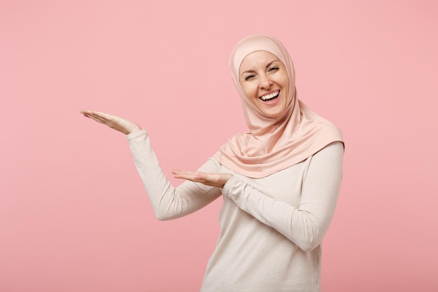 Enthousiaste souriante jeune femme musulmane arabe en vêtements légers hijab posant isolé sur fond rose en studio. Concept de style de vie de l'islam religieux des gens. Maquette de l'espace de copie. Pointant les mains de côté.