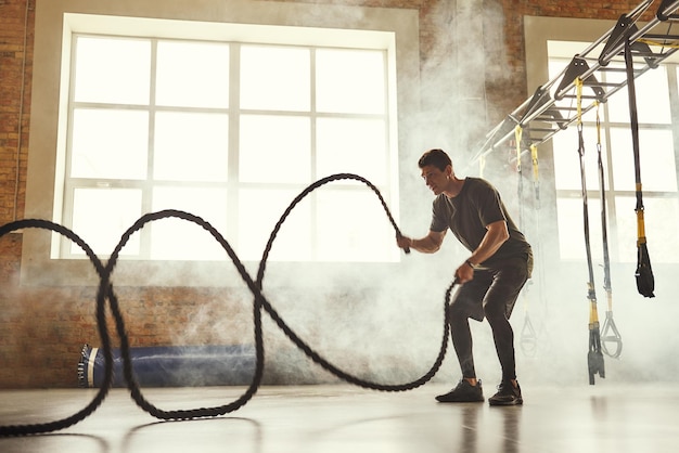 Entraînement CrossFit. Vue latérale d'un jeune homme athlétique avec un corps parfait faisant des exercices de crossfit avec une corde dans la salle de gym.