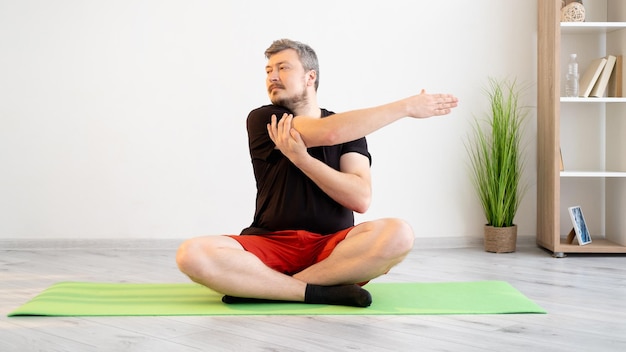 Entraînement à domicile Homme sportif Salle de fitness Mode de vie confiné Calme concentré en short de t-shirt assis pose de lotus faisant des exercices d'étirement pour les mains dans l'intérieur de la pièce lumineuse