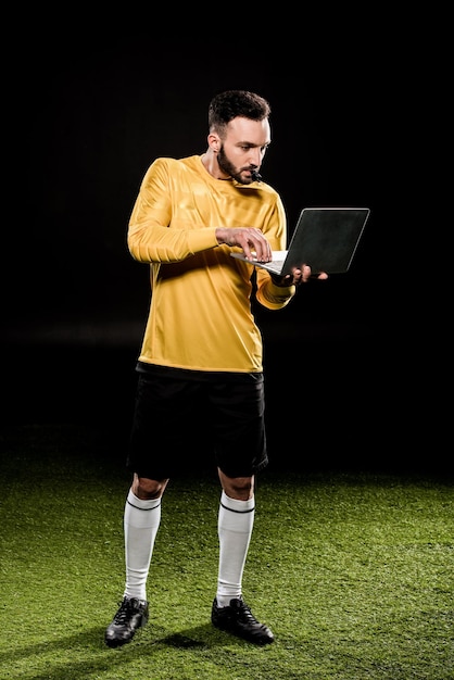 Entraîneur barbu regardant un ordinateur portable et sifflant isolé sur noir