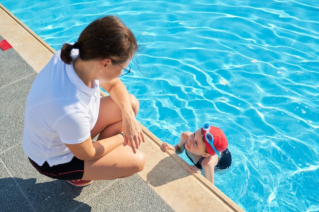 Entraîneur de femme parlant à la petite fille en chapeau de maillot de bain et lunettes près de la piscine extérieure