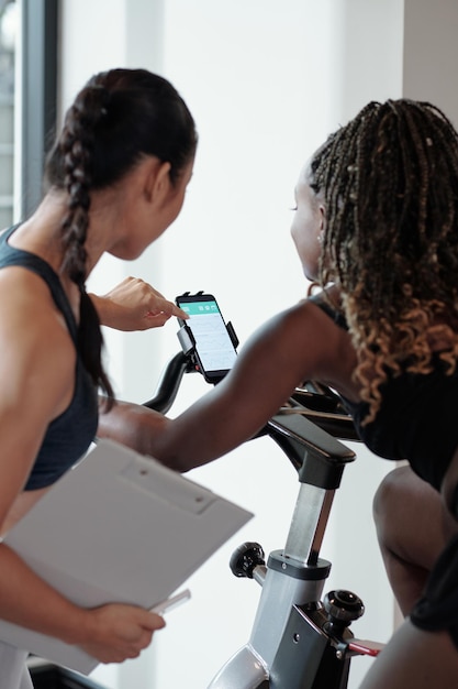 Photo entraîneur de fitness montrant au client comment contrôler la vitesse sur un vélo d'exercice dans une salle de sport