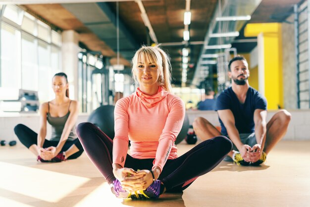 Entraîneur de fitness montrant un exercice de couple sportif pour les jambes qui s'étend en étant assis sur le sol de la salle de gym Dans le miroir de fond.