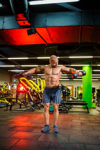 Un entraîneur masculin fort, adulte, en forme et musclé pose pour une séance photo dans une salle de sport sous les projecteurs en tenue de sport, montre ses muscles et tient des haltères avec confiance.