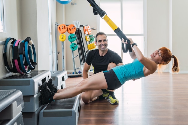 Entraîneur personnel à côté d'une fille faisant de l'exercice avec un trx tout en regardant la caméra en souriant dans une salle de sport.