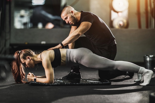 Entraîneur personnel regardant et aidant une jeune femme musclée à l'entraînement dans une salle de sport. Elle fait des exercices de planche pour son tronc.