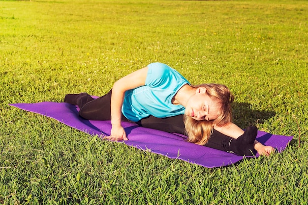 Une entraîneure blonde dans un haut court sportif et des leggings de gym fait une large ficelle sur le tapis pour le yoga les mains sont levées vers le haut sur le tapis de yoga un jour d'été