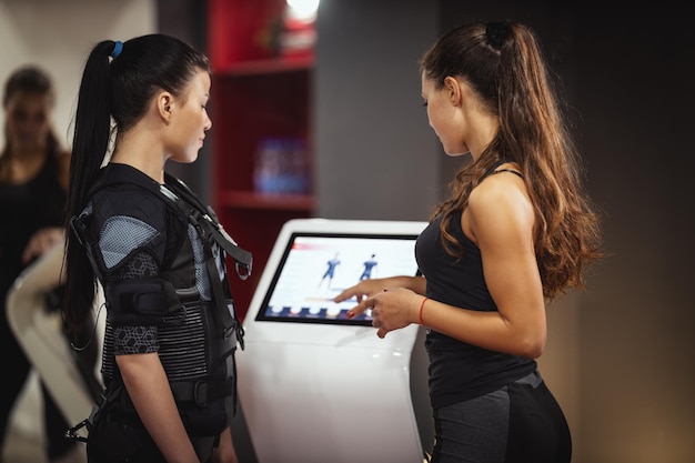 Une entraîneuse personnelle enseigne à une jeune femme souriante à s'entraîner avec un appareil EMS dans la salle de sport.