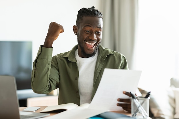 Entrepreneur afro-américain excité célébrant le succès tenant des papiers et souriant joyeusement levant la main