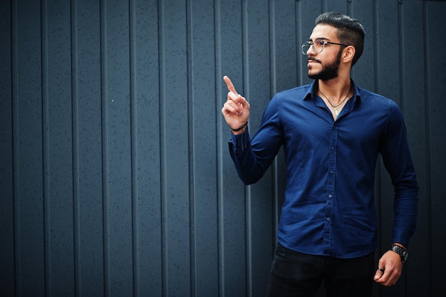 Un entrepreneur du Moyen-Orient porte une chemise bleue, des lunettes, contre un mur d'acier, montre le doigt vers le haut.