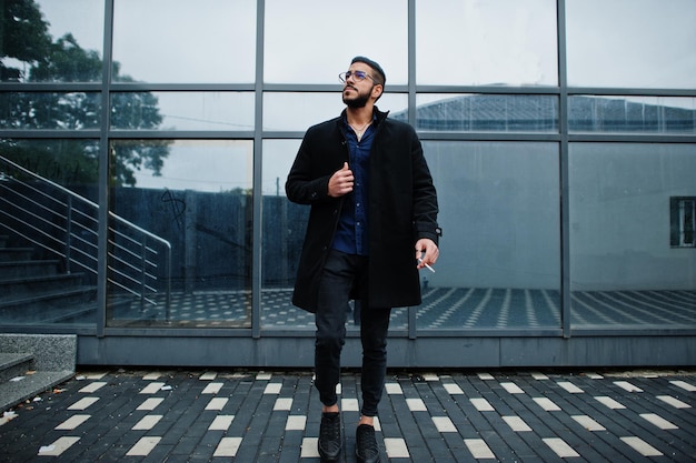 Un entrepreneur du Moyen-Orient porte un manteau noir et une chemise bleue, des lunettes contre une cigarette dans un immeuble de bureaux.