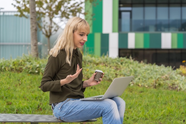 Entrepreneur de fille blonde caucasienne dans le parc d'affaires effectuant une vidéoconférence avec un café à emporter. Polygone moderne parmi la nature