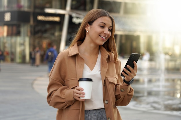 Entrepreneur jeune femme occupée Application Smartphone Employé de bureau à l'extérieur Tasse à café Cappuccino Centre commercial Architecture fond urbain moderne