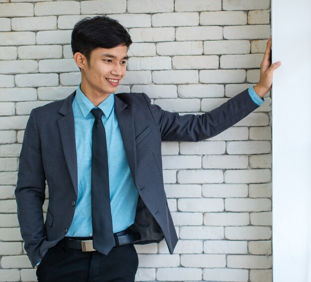 Entrepreneur masculin asiatique confiant en costume souriant et regardant ailleurs tout en s'appuyant sur un mur de briques