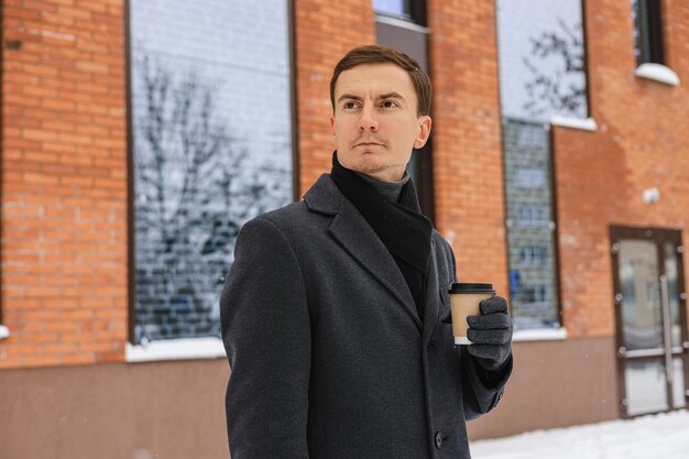 Entrepreneur masculin prospère avec une tasse de café en papier pour se tenir debout dans la rue de la ville en hiver