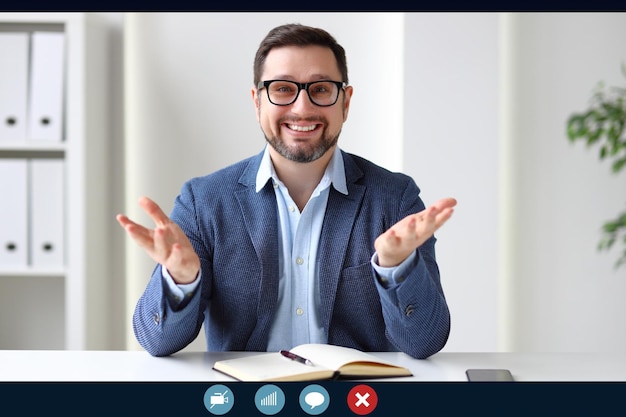 Entrepreneur masculin ravi souriant pour la caméra et gesticulant tout en faisant un appel vidéo pendant le travail dans un bureau moderne