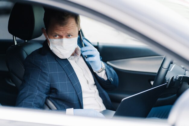 Un entrepreneur prospère et sérieux pose dans son propre transport, a une conversation téléphonique, utilise des technologies modernes pour être toujours en contact, porte un masque médical et des gants pendant la quarantaine et la pandémie