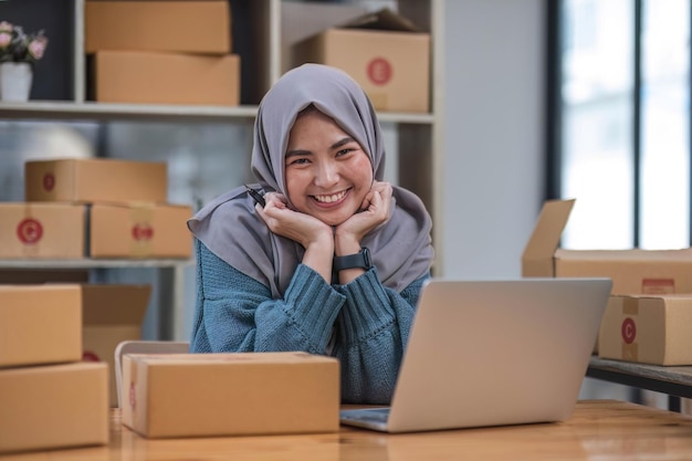 Photo entrepreneure indépendante asiatique musulmane sourire pour le succès des ventes après avoir vérifié la commande d'une boutique en ligne dans un ordinateur portable au bureau à domicile concept d'entreprise marchande en ligne et de commerce électronique