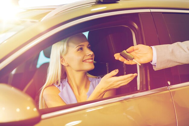 Entreprise automobile, vente de voitures, consommation et concept de personnes - femme heureuse prenant la clé de voiture du concessionnaire dans le salon de l'auto ou le salon