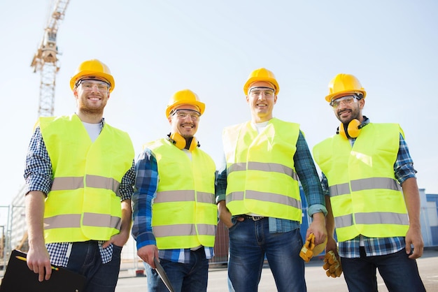 entreprise, bâtiment, travail d'équipe, technologie et concept de personnes - groupe de constructeurs souriants en casque avec ordinateur tablette et clipboard à l'extérieur