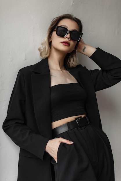 Entreprise à la mode jeune belle femme modèle avec des lunettes de soleil à la mode en noir élégant haut blazer et pantalon se tient près d'un mur blanc dans la rue