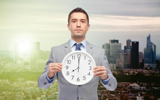 entreprise, personnes, décalage horaire et concept de travail - homme d'affaires tenant une horloge indiquant 8 heures au lever et au coucher du soleil sur fond de ville