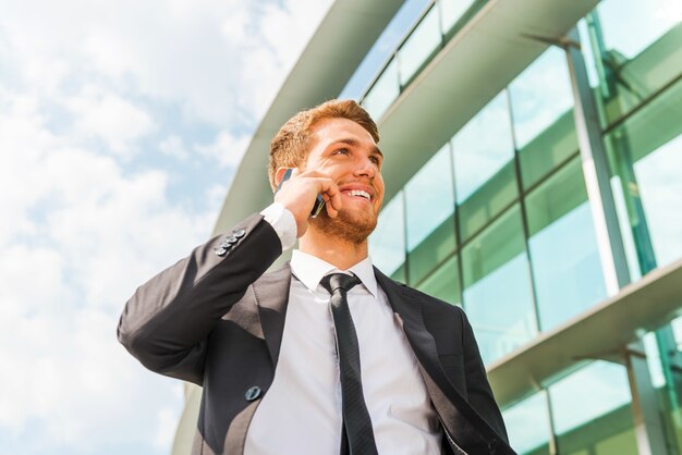 Photo entretien d'affaires confiant. jeune homme gai en tenue de soirée parlant au téléphone portable et regardant ailleurs tout en se tenant à l'extérieur et contre la structure du bâtiment