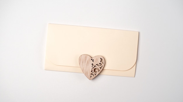 Une enveloppe avec un cœur en bois dessus sur fond clair (blanc). Mise à plat, vue de dessus