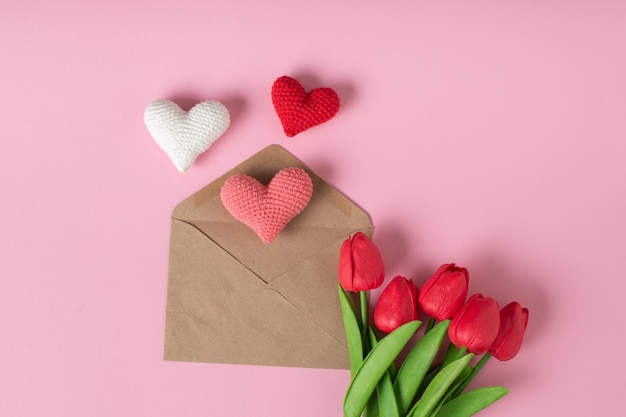 Une enveloppe kraft avec trois coeurs tricotés et un bouquet de tulipes sur fond rose