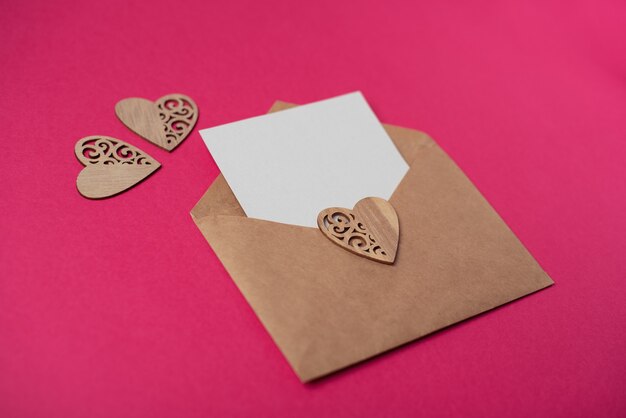 Enveloppe de métier avec une feuille vierge et des coeurs en bois sur le fond rose chaud.