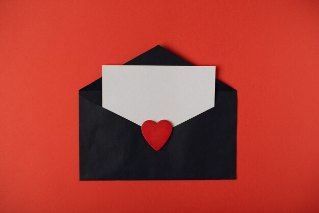 Enveloppe noire avec une feuille vierge à l'intérieur et coeur en bois rouge sur fond rouge. Concept de la Saint-Valentin. Mise à plat, vue de dessus.