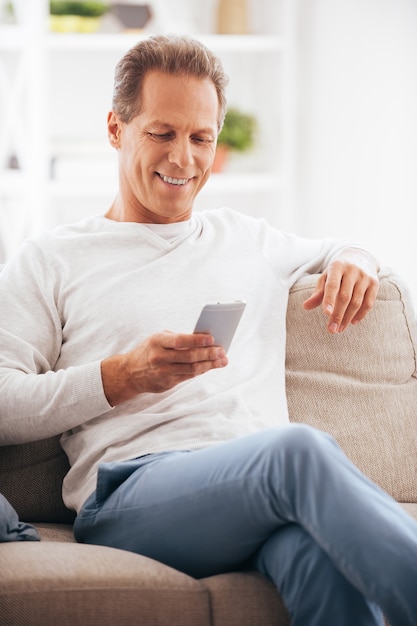 Envoyer un SMS à un ami. Homme mûr confiant tenant un téléphone portable et le regardant avec le sourire alors qu'il était assis sur le canapé à la maison